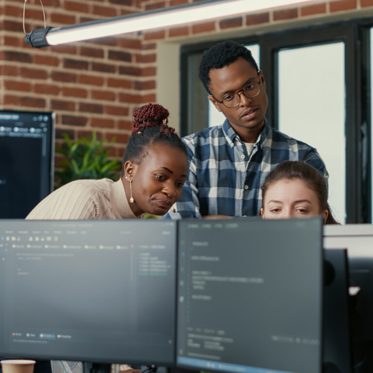 Developers brainstorming ideas looking at code on computer screens asking feedback from senior developer while intern joins the discussion. Junior programmers collaborating on group project.